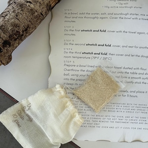 Könnte beinhalten: Ein Rezept für Sauerteigbrot mit Zutaten und Anweisungen. Ein kleiner Leinenbeutel mit der Aufschrift "Dry Sourdough Starter 10 grams" liegt neben einem kleinen Haufen Anstellgut. Ein Stück Holz befindet sich in der oberen linken Ecke.