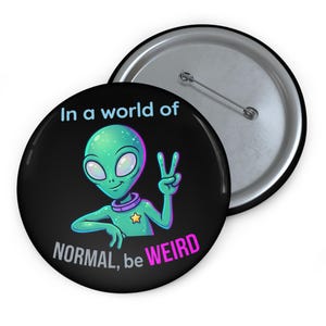 Peut inclure: Un badge rond avec une épingle au dos, présentant un fond noir et un personnage extraterrestre vert faisant un signe de paix. Le texte In a world of NORMAL, be WEIRD est écrit en bleu clair et rose. Le dos montre un mécanisme d'épingle en métal.