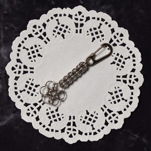 May include: A silver-colored keychain with a flower-shaped charm and a chain link design. The keychain is attached to a silver carabiner clip. The keychain rests on a white, lacy paper doily, set against a dark, marbled background.