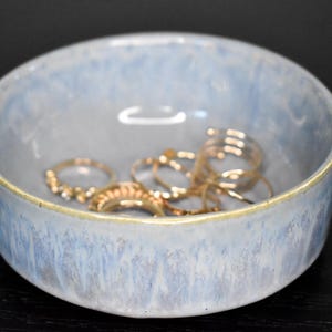 May include: A small, round ceramic bowl with a mottled light blue and white glaze finish. The rim features a thin, brownish-gold accent line. Inside the bowl, there is a collection of several gold-toned metal rings of varying designs.