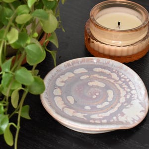 May include: A round ceramic trinket dish featuring a swirling pattern of soft lavender, white, and pale pink glazes. Beside the dish sits a small, clear glass jar containing a cream-colored candle with a visible black wick.
