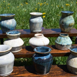 May include: A collection of handmade ceramic vases in various shapes and sizes. The vases feature a range of colors, including blue, white, and green, with unique glaze finishes. Displayed on a wooden shelf against a backdrop of green grass and yellow flowers.