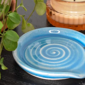 May include: A round, light blue ceramic spoon rest featuring a white spiral pattern in the center. The dish has a smooth, glossy finish with a slightly raised edge and a small indentation on one side, designed to hold a kitchen utensil securely.