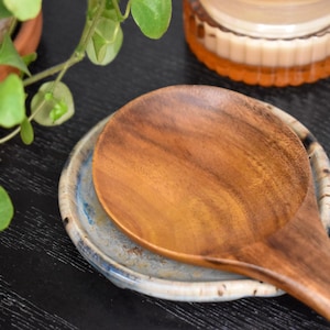 May include: A smooth, polished wooden spoon resting on a small, round, handmade ceramic spoon rest. The spoon rest features a mottled blue and white glaze with a textured edge, while the wooden spoon displays a rich, natural grain pattern.