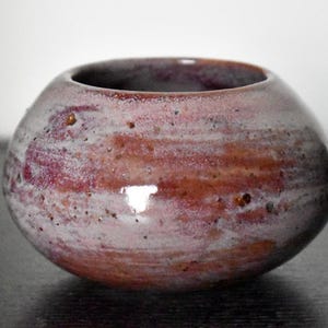 May include: A small, round ceramic bowl with a wide, open top. The exterior features a mottled glaze with blended shades of dusty pink, deep purple, and earthy brown, accented by small, dark speckles and a smooth, glossy finish throughout.
