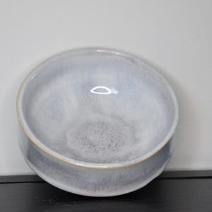 May include: A round ceramic bowl with a smooth, glazed finish in shades of light blue, white, and grey. The interior features a speckled texture at the base, while the exterior rim shows a subtle, thin line of natural clay color around the edge.