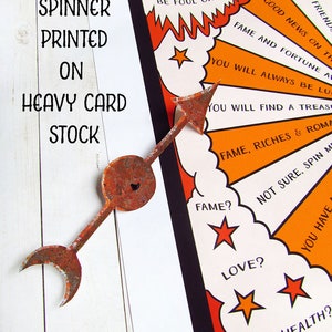 May include: A vintage-style fortune teller wheel printed on heavy card stock. The wheel has a rusty metal spinner with an arrow. The wheel has a colourful design with orange, red, black, and white colours. The wheel has text that reads "Wheel of Fortune" and "Spin the wheel if you dare, will your fortune be foul or fair?"