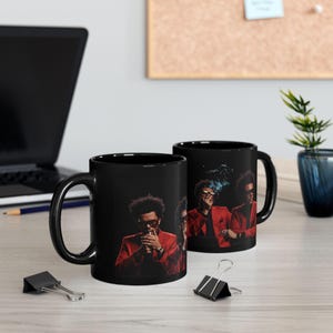 May include: Two black ceramic coffee mugs featuring a portrait of a person in a red suit. The person is smoking a cigarette. The mugs are on a light-colored surface with a laptop and office supplies in the background.