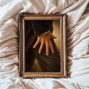 May include: A framed painting featuring a close-up of a person's hand resting on their leg. The painting has a gold-colored ornate frame. The person in the painting is wearing a dark suit. The background is a light tan color.