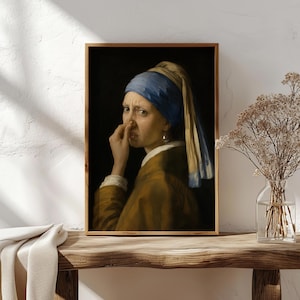 May include: A framed wall art print depicting a person wearing a blue headscarf and a brown jacket, holding their nose with one hand. The figure has a pearl earring and is set against a dark background, presented in a simple wooden frame.