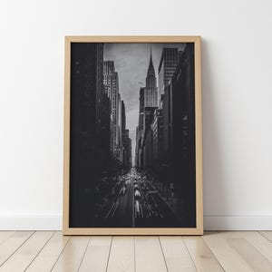 May include: Black and white framed print of a New York City street scene. The image captures a perspective view down a city street lined with tall buildings, with the Chrysler Building visible in the distance. The print is in a light wood frame.
