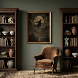 May include: A framed wall art print depicting a witch flying on a broomstick against a full moon, positioned between two dark wood bookcases filled with books, ceramic vases, a human skull, and a torso sculpture, with a brown velvet armchair.