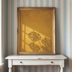 May include: A rectangular wall art piece featuring a solid mustard yellow background with a dark shadow of monstera leaves and a window pane cast across the surface, enclosed in an ornate, metallic gold-toned frame with decorative detailing.