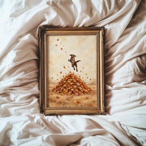 May include: A rectangular framed print featuring a dark-colored dog jumping into a large pile of orange and brown autumn leaves. The scene is set in a field with scattered foliage against a soft, light-colored background, all encased in a gold frame.