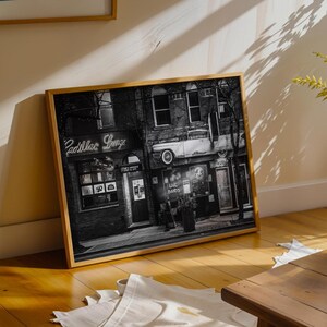 May include: Black and white framed print of a vintage building with a classic car mounted on the side. The sign reads "Cadillac Lounge" in neon. The image is displayed on a wooden floor.