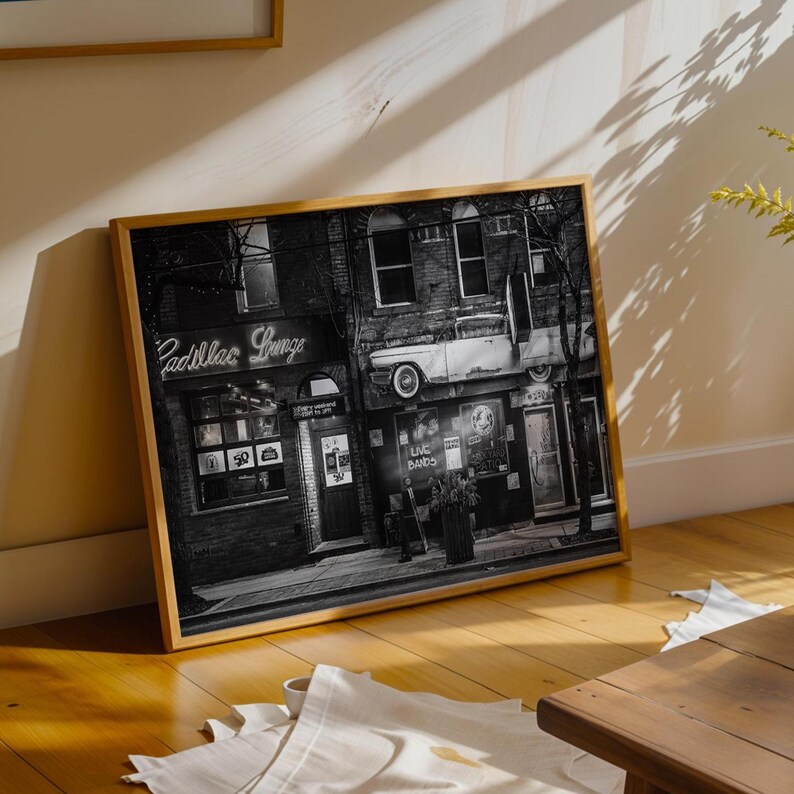 May include: Black and white framed print of a vintage building with a classic car mounted on the side. The sign reads "Cadillac Lounge" in neon. The image is displayed on a wooden floor.