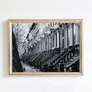 May include: Black and white framed print of a row of brownstone buildings. The image captures the architectural details of the buildings, including the steps, railings, and decorative columns. The photograph is in a gold-colored frame.