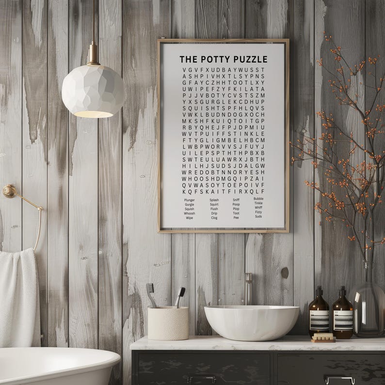 May include: A framed word search puzzle titled "The Potty Puzzle" hangs on a wooden wall. The bathroom scene includes a white sink, a decorative light fixture, and a vase with branches. The puzzle's words are related to bathroom activities.