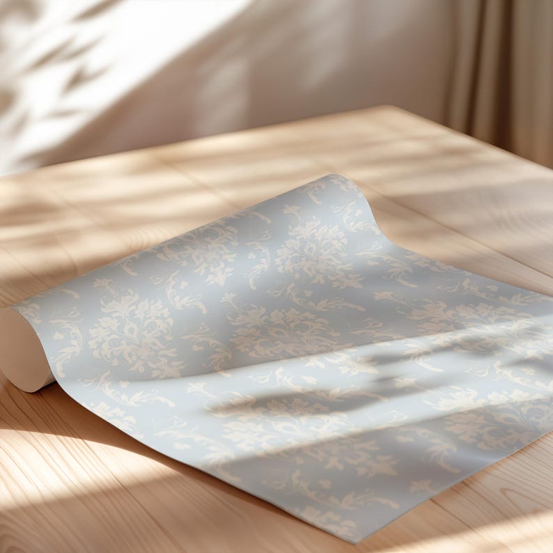 May include: A roll of wallpaper featuring a light blue background with a repeating white damask pattern. The paper is partially unrolled, displaying the intricate floral and scrollwork design across its smooth, matte surface texture.