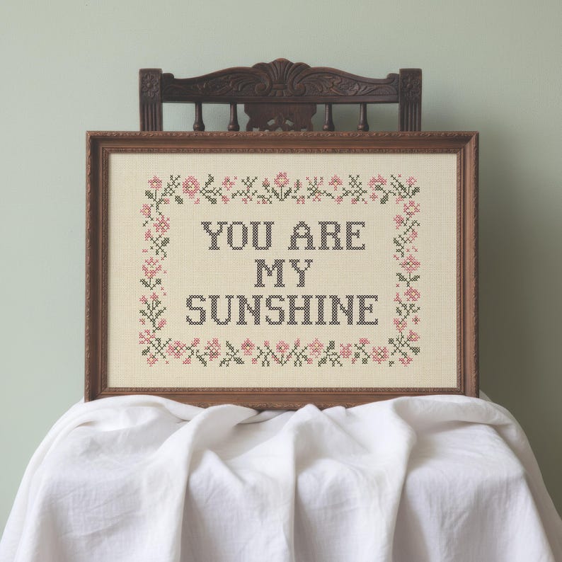 May include: A framed cross-stitch with the words "YOU ARE MY SUNSHINE" in black thread. The design is surrounded by a border of pink and green floral elements. The frame is a warm brown color, and the piece is displayed on a white cloth.