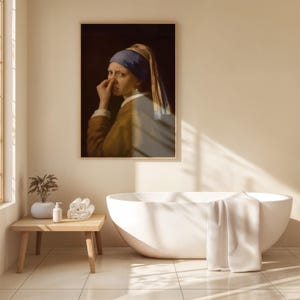 May include: A framed wall art print depicting a person wearing a blue headscarf and a brown garment, holding their nose with one hand. The artwork is displayed on a beige wall above a white freestanding bathtub and a small wooden side table.