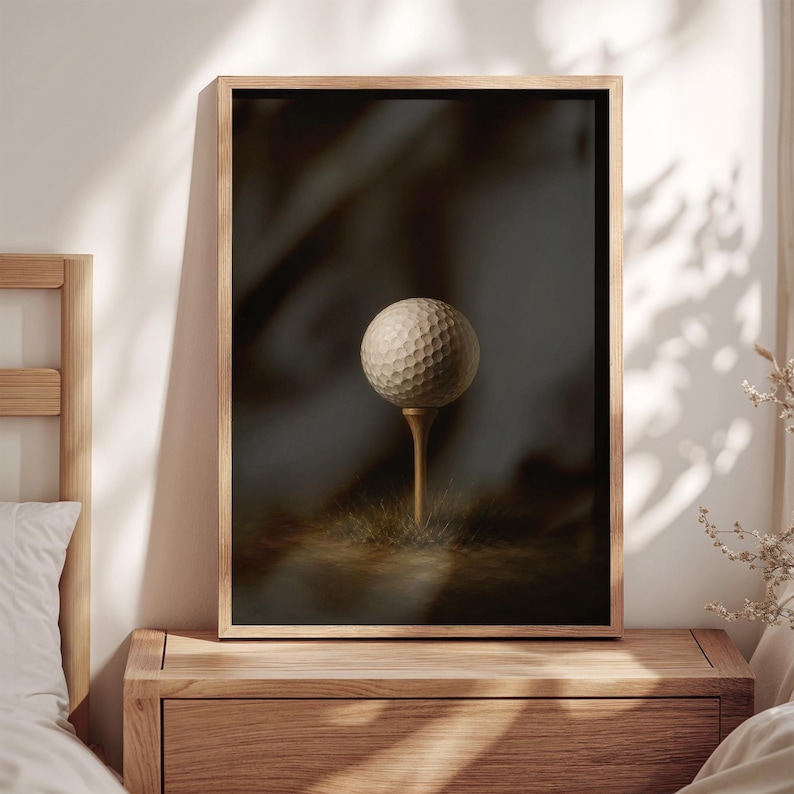 Op de afbeelding: Een ingelijste wandkunstprint met een witte golfbal met putjes op een houten tee. De golfbal is centraal geplaatst tegen een donkere, sfeervolle achtergrond met zachte verlichting, wat zorgt voor een gefocuste compositie van een golfbal.