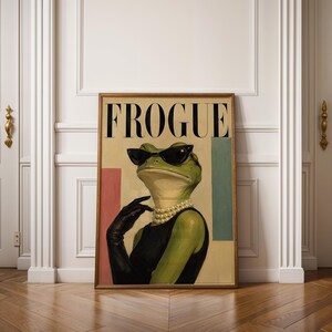 May include: A framed wall art print titled FROGUE featuring a green frog wearing black cat-eye sunglasses, a multi-strand pearl necklace, and a black sleeveless dress with a matching black glove. The background includes pink and teal rectangles.