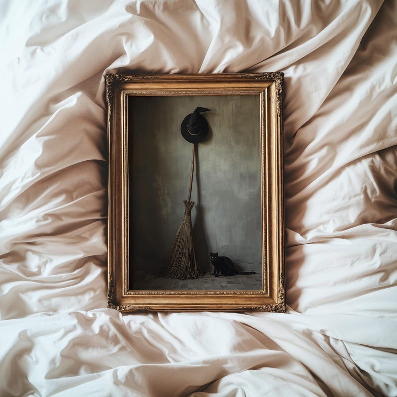 May include: A rectangular gold-toned ornate frame containing a print of a black pointed hat hanging above a straw broom, with a small black cat sitting on the floor against a muted green wall, all presented in a vertical composition.