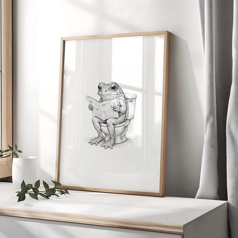 Puede incluir: Ilustraci&oacute;n en blanco y negro enmarcada de una rana leyendo un peri&oacute;dico en un inodoro. La obra de arte se presenta en un marco de madera clara. La imagen es una representaci&oacute;n humor&iacute;stica de la vida cotidiana.