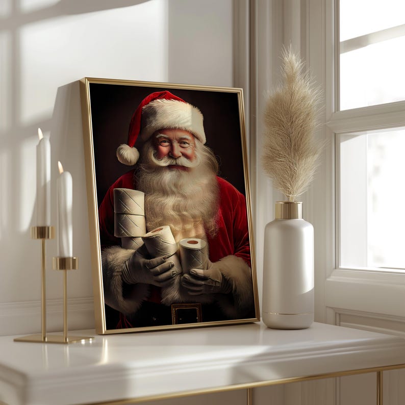 May include: A framed print featuring Santa Claus holding rolls of toilet paper. Santa is wearing a red suit and hat with white trim, and has a long white beard and mustache. The print is in a gold frame and sits on a white surface.