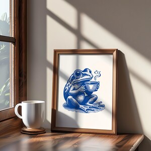 May include: A framed illustration of a blue frog sitting and holding a cup of coffee. The frog is in a relaxed pose with its eyes closed. The illustration is on a white background with a brown frame.
