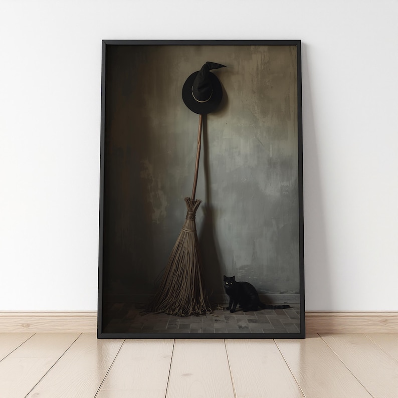 May include: A framed wall art print featuring a black pointed hat hanging above a long-handled wooden broom with straw bristles. A small black cat sits on the floor in front of the broom. The scene is set against a muted, textured grey wall.