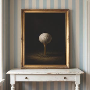Op de afbeelding: Een ingelijst kunstwerk aan de muur met een witte golfbal op een houten tee, tegen een donkere, effen achtergrond. Het kunstwerk is gevat in een sierlijke, goudkleurige lijst, wat het contrast tussen de bal en de donkere ruimte benadrukt.