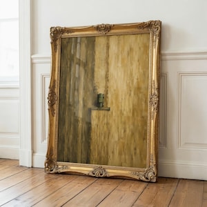 May include: A large rectangular painting in an ornate gold frame with intricate carved details. The artwork features a vertical composition with shades of ochre, brown, and tan, depicting a small green object resting on a horizontal ledge.