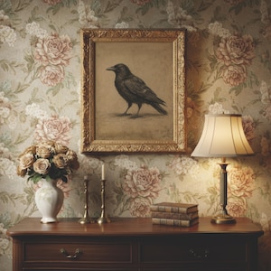 May include: A framed charcoal drawing of a black crow hangs above a wooden dresser. The dresser holds a white vase with beige roses, two brass candlesticks, a stack of three leather-bound books, and a table lamp with a cream-colored shade.