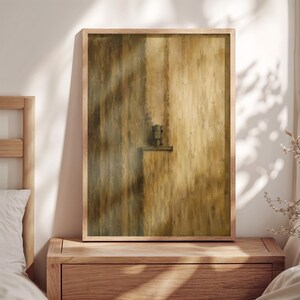 May include: A rectangular wall art print featuring a vertical abstract composition in shades of ochre, gold, and dark brown. The artwork displays a textured, brushstroke-heavy surface with a small, dark, centered geometric element resembling a latch.