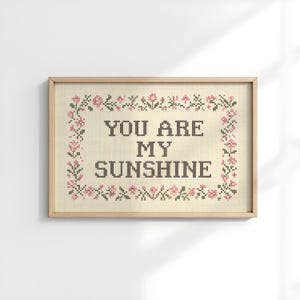 May include: Cross-stitch artwork in a light wooden frame. The artwork features the text "YOU ARE MY SUNSHINE" in black thread, surrounded by a floral border with pink and green accents. The background is a light beige fabric.