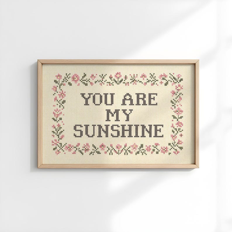 May include: Cross-stitch artwork in a light wooden frame. The artwork features the text "YOU ARE MY SUNSHINE" in black thread, surrounded by a floral border with pink and green accents. The background is a light beige fabric.