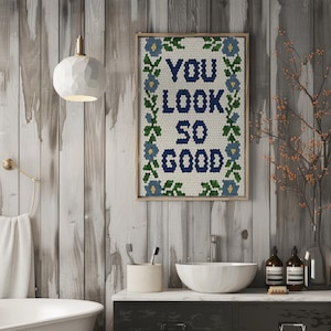 Puede incluir: Una obra de arte enmarcada con el texto "YOU LOOK SO GOOD" en azul marino, rodeada por un borde floral. La obra de arte se exhibe en un ba&ntilde;o con un lavabo blanco, una toalla y una l&aacute;mpara decorativa.