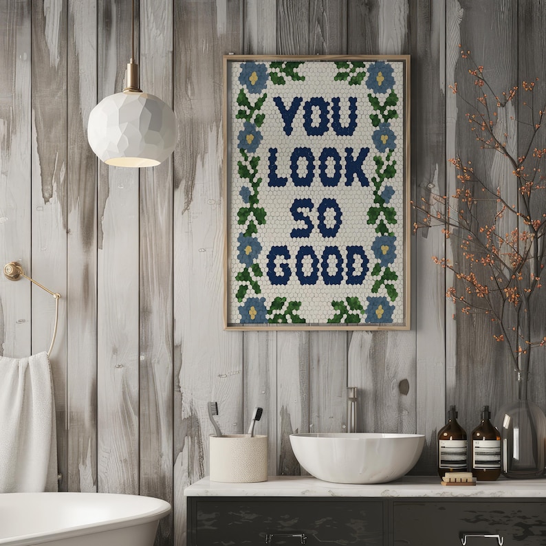 Puede incluir: Una obra de arte enmarcada con el texto "YOU LOOK SO GOOD" en azul marino, rodeada por un borde floral. La obra de arte se exhibe en un ba&ntilde;o con un lavabo blanco, una toalla y una l&aacute;mpara decorativa.