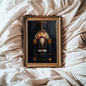 May include: A framed painting of a nutcracker soldier wearing a dark blue uniform with gold trim, a tall hat, and a white beard. The figure has a moustache, rosy cheeks, and stands against a dark background. The frame is ornate and metallic gold.