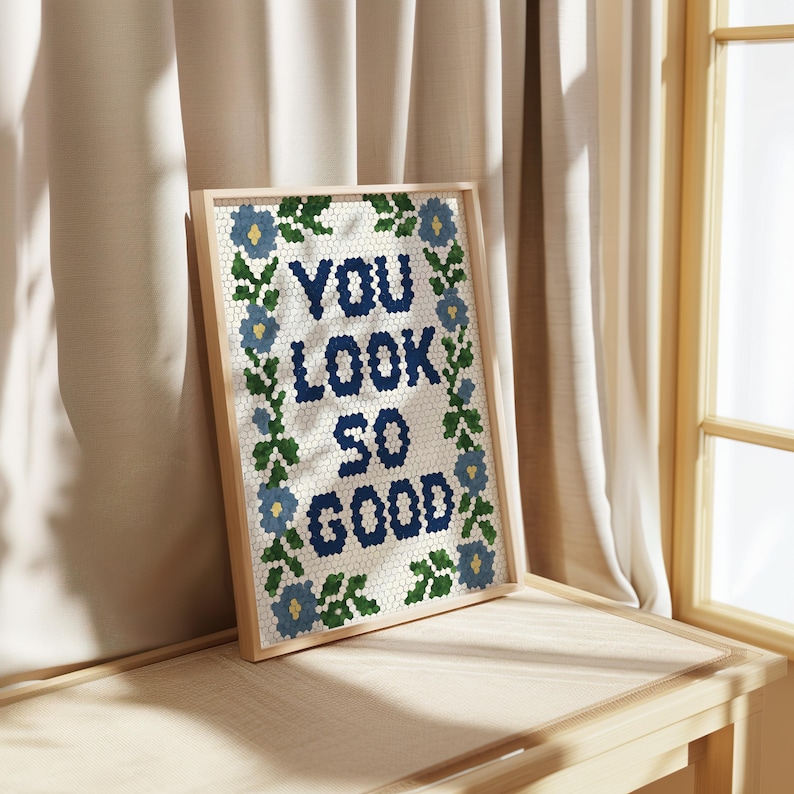 Puede incluir: Una obra de arte enmarcada con el texto "YOU LOOK SO GOOD" en azul marino, rodeada de motivos florales azules y hojas verdes. La obra est&aacute; sobre un fondo blanco con un patr&oacute;n de panal. El marco es de madera clara.