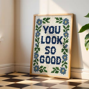 Puede incluir: Un cuadro enmarcado con la frase "YOU LOOK SO GOOD" en azul marino, rodeado por un borde floral azul y verde. La obra de arte tiene un efecto de mosaico y est&aacute; enmarcada en un marco de madera clara.