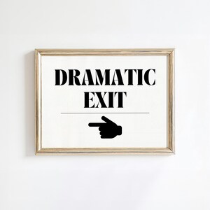 May include: A framed sign with the words "DRAMATIC EXIT" in bold black letters. Below the text is a horizontal line, and a black hand pointing to the right. The frame is a light gold color.