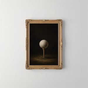 Op de afbeelding: Een witte golfbal staat op een houten tee in een plukje gras, gecentreerd tegen een donkere achtergrond. Het geheel is gevat in een sierlijke, rechthoekige goudkleurige lijst met gedetailleerd houtsnijwerk langs de randen.