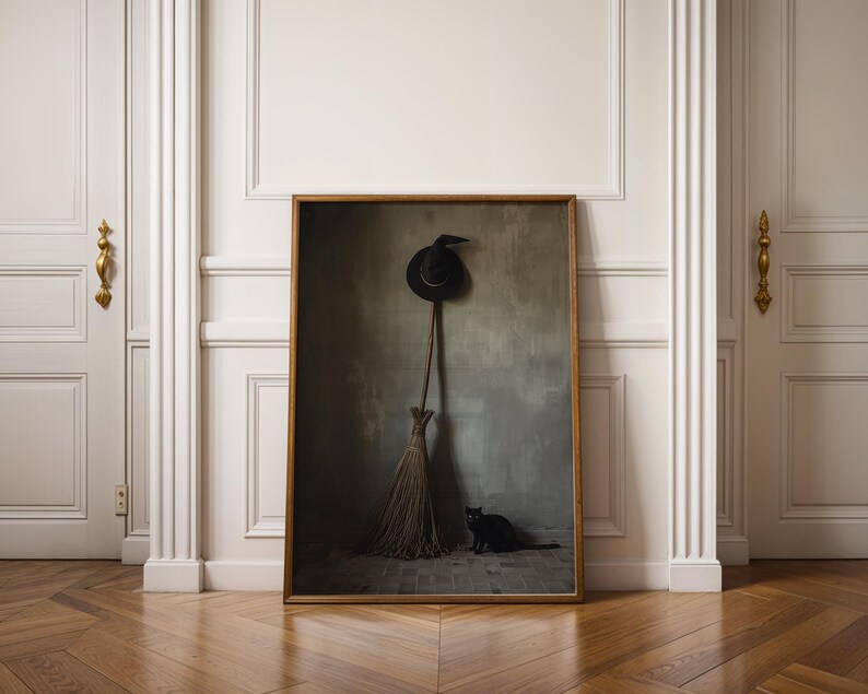 May include: A framed rectangular wall art print featuring a dark, moody scene with a black pointed hat hanging above a wooden broomstick, and a small black cat sitting on a stone floor against a textured grey wall in a dimly lit room.