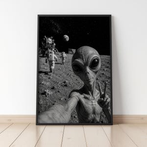 May include: A black and white framed wall art print depicting an alien in the foreground taking a selfie on the moon surface. In the background, two astronauts in space suits walk toward a lunar lander near an American flag under a starry sky.