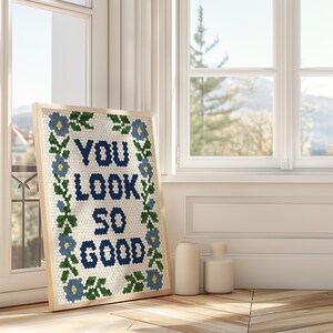 Puede incluir: Una obra de arte enmarcada con el texto "YOU LOOK SO GOOD" en azul oscuro, rodeada por un borde floral. La obra de arte se exhibe en un suelo de madera cerca de una ventana. Dos jarrones blancos est&aacute;n junto a la obra.
