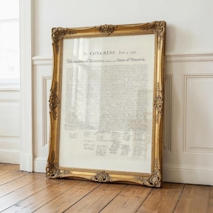 May include: A framed document, the Declaration of Independence, is displayed. The gold frame has ornate detailing. The document's text is visible, with signatures at the bottom. The frame leans against a white wall and rests on a wooden floor.