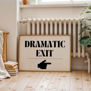 Puede incluir: Un letrero enmarcado con las palabras "DRAMATIC EXIT" en letras negras y una mano negra que señala hacia la derecha. El letrero está en una habitación con un radiador, una planta y un suelo de madera.